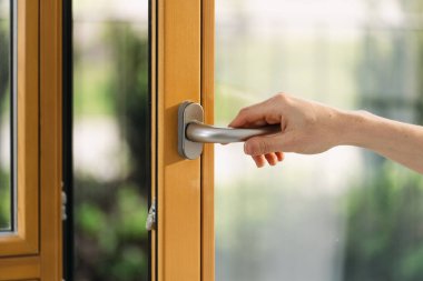 Kesilmiş bir kadın çift camlı pencerede metal sapı çekiyor. Dairesinde kadın açık plastik balkon kapısı var.