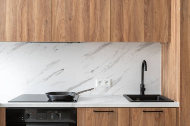 Beyaz tezgahlı mutfak, siyah lavabo, musluk ve sobanın üstü. Beyaz mermer kiremitler ve ahşap dolaplarla iç mekan. İndüksiyon ocağında kızartma tavası. Modern ev aletleriyle dolu bir daire.