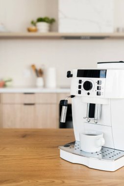 Tahta tezgahın üzerinde beyaz espresso makinesinin dikey görüntüsü ve damlama tepsisinde bir fincan kahve. Mutfak gereçleri bulanık arkaplana karşı masada, kopyalama alanı
