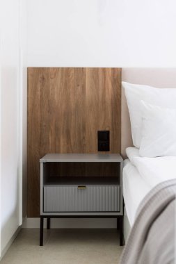 Çağdaş bir yatak odasında ahşap bir duvar paneli bulunan, düzgün yapılmış bir yatağın yanındaki çekmecesi olan bir komodin. Konforlu bir dairede ya da otel süitinde modern iç mekan unsuru. Odada komodin var.