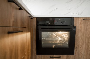 Evde yemek pişirmek için ev aletleri olan modern bir daire. Mutfaktaki ahşap dolapta açık kapı ve ışık bulunan siyah fırının önü.