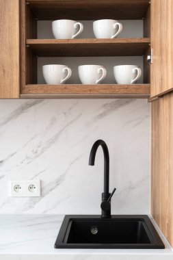 Modern iç tasarımın elementleri. Siyah mutfak lavabosunun dikey çekimi, ahşap dolapların altında su musluğu var. Duvarda asılı ahşap dolap ve içinde beyaz kupalar.