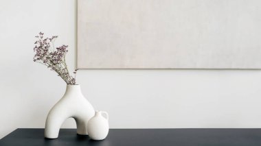 Pürüzsüz, beyaz bir vazo, karanlık bir masanın üzerinde, tarafsız bir arkaplana karşı kurumuş çiçekleri tutar. Minimalist bir estetik sergiler. Rafta ev dekorasyonu olan oturma odası ve duvarda resim çerçevesi kopyala