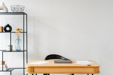 Ahşap masa üzerinde iş kağıtları ve laptop olan çağdaş bir ofis çalışma alanı. Ofis masasının arkasındaki siyah sandalye, dekoratif eşyalarla süslenmiş raf ünitesine yakın. Eğitim için ortak çalışma alanı