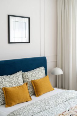 Huzurlu bir yatak odası, yumuşak beyaz perdeler ve duvar dekorunun altında fotokopi sanatıyla tamamlanmış, desenli sarı yastıklı mavi bir yatak başlığına sahiptir.