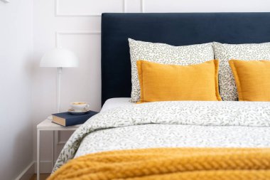 Parlak yatak odasında yumuşak bir lacivert yatak başlığı, çiçek desenli yatak örtüsü ve yanında okuma lambası, kitap ve bir fincan kahve ile tamamlanmış yastıklı sarı dekoratif yastıklar sergileniyor.