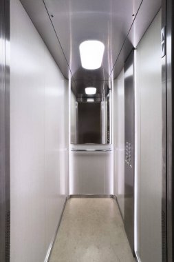 Çok katlı bir binada boş yolcu asansörü, dikey çekim. Modern asansör içinde metal duvarlar, ayna ve kabinin içinde parlak ışıklandırma sergileniyor.