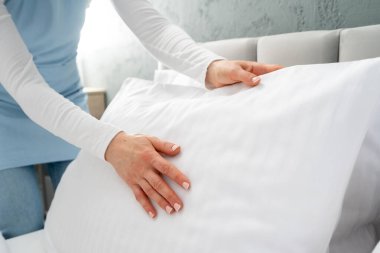Mavi üniforma giyen birinin otel odasında bir yatak yaptığı yakın plan görüntüsü. Kadın yastığı yastığın yanına, beyaz ve taze yastık kılıfına koyar. Hizmetçi misafir için yatak odasını hazırlıyor.