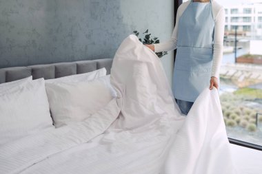 Otel odasındaki yatağı mavi önlüklü kadın yapıyor. Oda servisi, ev işleri ve çamaşır işleri kavramı. Bahar temizliği ve çarşaf değişimi. Beyaz yorgan örtülü battaniye.
