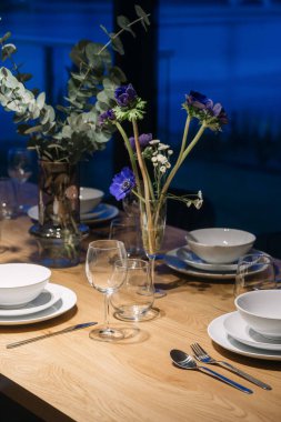 Akşam yemeği için beyaz tabak, gümüş çatal bıçak ve şarap kadehleriyle dikey çekim. Masanın ortasındaki vazoda duran mavi çiçekler..