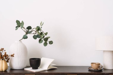 Kitap, okuma lambası, bir fincan kahve, zarif kişisel aksesuarlar ve okaliptüslü vazo, şık bir oturma odasında. Arkaplanda beyaz kopya alanı duvarına karşı ev dekorasyonu