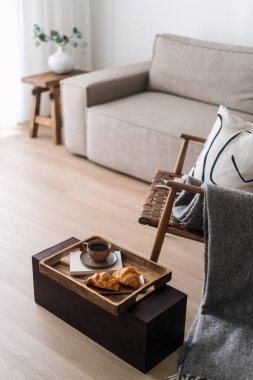 Kahve fincanı ve ahşap kahvaltı tepsisinde taze hamur işi. Konforlu evde sabah çayı ve kruvasan. Otel ve oda servisi konseptleri. Koltuk ve modüler kanepe tasarlanmış rahat bir dairede.