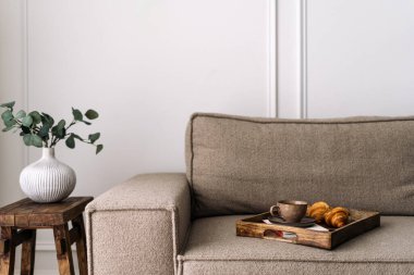 Kahvaltı tepsisiyle rahat kanepe ev dekorlu yan masaya yakın. Kahve fincanı ve kruvasanla birlikte beyaz seramik vazoda yeşil okaliptüs bitkisi. Konforlu yaşam, otel odası servisi kavramları.