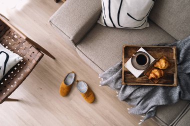 Dekoratif yastıklı rahat kanepe ve ahşap tepside kruvasan ile kahve fincanı. Kahvaltıyı evde yaparız. Oturma odasının tam üstünde. Yüksek açılı kanepe rattan koltuğa yakın.