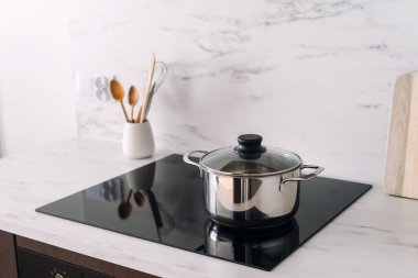 Bulanık arka plandaki mermer mutfak masasına karşı cam seramik kapaklı paslanmaz çelik tavaya odaklan. Modern indüksiyon sobasında evde yemek pişirmek. Fırında metal mutfak gereçleri