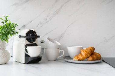 İçinde espresso makinesi olan mermer mutfak tezgahı, bir tabak altın çıtır kruvasan ve bir yığın beyaz bardak. Kahve makinesi pasta ve fincanlara yakın.