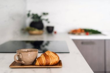 Beyaz mermer tezgahta kruvasan ve kahve fincanı. Yemek ve içecek mutfak tezgahında. Fransız pastasına ve seramik bardakta bitki çayına odaklan. Arka planda bulanıklık olan yemek masasında.