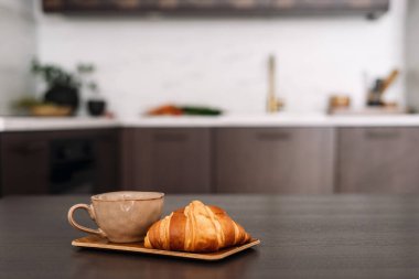 Taze kruvasan ve ahşap masanın üzerindeki kahve fincanı ile yüzeyinde fotokopi alanı olan bulanık mutfak arka planına odaklan. İç kısımlarda hala hayatın detayları var. Hafta sonunun tadını çıkar