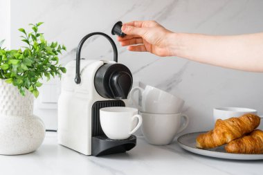 Kadın kahve makinesine kahve kapsülü sokuyor. Kahve makinesi, fincanlar ve kruvasanlar mutfak tezgahında mermer kaplamalı. Evdeki ev aletleri.