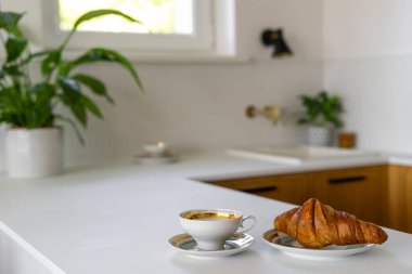 Temiz, minimalist mutfak tezgahında arka planda bitkiler olan taze bir Fransız kruvasanının yanında sabah kahvesi.