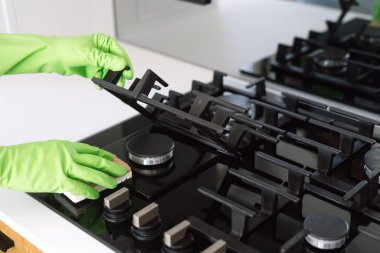 Yeşil lastik eldiven giyen biri, modern bir mutfakta etkin temizlik için çıkarılabilir ızgaraları dikkatlice tutarken gaz sobasını süngerle temizler.