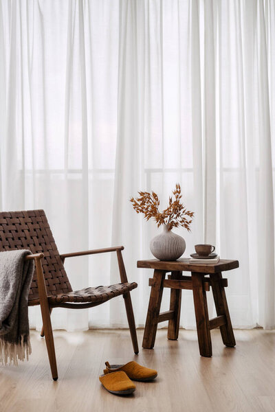 Minimalist interior with plaid on wicker chair, coffee cup and home decor on side table and slippers on wooden floor in room. Simple living concepts. Hygge and cozy space in comfort apartment