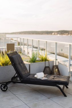 Siyah şezlongla ilgili açık kitap, dikey çekim. Çatıdaki verandada su manzaralı gri yastıklı şezlong. Otel, yaz tatili, dinlenme ve dinlenme konseptleri.