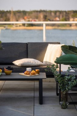 Çatının terasındaki gri kanepeye karşı gevrek kruvasan ve kahve fincanı. Modern bahçe mobilyalarıyla balkon. Dinlenme bölgesi ve bir su kütlesi manzaralı konut binası, dikey çekim