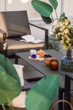 Teras ile sıcak bir hafta sonu. Üzerinde kruvasan, meyve, kahve fincanı ve mum olan gri veranda masasına odaklan. Balkonda öğle yemeği. Açık hava mobilyası. Sandalyede kitap