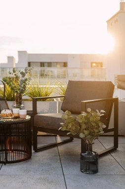 Şehir manzaralı, balkondaki yan masanın yanındaki gri sandalyenin dikey görüntüsü. Teraslı bir daire, teraslı mobilyalar, yemek, vazodaki bitkiler ve çiçekler. Sıcak güneş ışığı rahatlatıcı bir atmosfer yaratıyor.