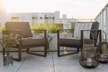 Açık balkonlu, koltuklarda dekoratif yastıklı, mumlu fenerlere yakın bir ev. Çatıdaki terasta arka planda şehir manzarası olan iki modern sandalye.