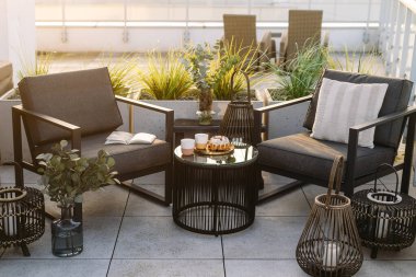 Balkonda gece fenerlerine yakın iki modern teras sandalyesi. Çatıdaki cam masanın yanındaki koltuklarda yeşil saksı bitkileri var. Arka planda şehir manzaralı konut binası