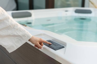 Arka bahçedeki terasta lüks jakuzi. Kadın modern banyoda su sıcaklığını seçer, arka plan ışıklarını seçer, hidromasaj jetini çalıştırır.