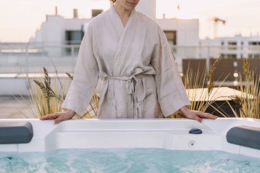 Bornozlu kadın jakuzinin yanında durup yazın otelin çatısında günbatımının tadını çıkarıyor. Sabah güneşinde mavi filtreli suyla modern küvetin yanında bir kadın. Çatı katında banyosu olan bir teras.