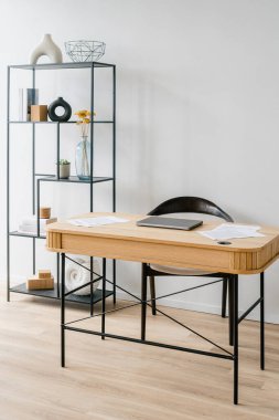 Çalışma alanının dikey görüntüsü dizüstü bilgisayar ve dokümanlar ahşap bir masa üzerinde, siyah sandalye ve ev ofisinde dekoratif eşya ve kitapları gösteren bir raf ünitesi