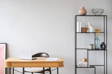 Tahta masadaki dizüstü bilgisayar tarafsız bir odada dekoratif eşyalarla dolu siyah bir rafın yanında duruyor. Eğitim için merkez ofis. Yaşam alanındaki gri duvara karşı masa