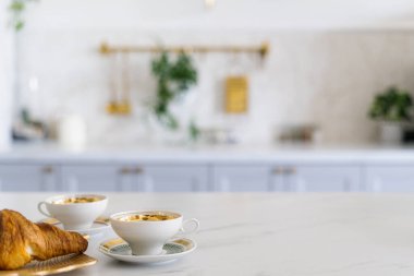 Parlak, şık bir yemek odasında iki fincan kahve ve mermer tezgahın üzerinde bir kruvasan. Mutfak dolaplarının arka planında yiyecek ve içeceklerle dolu beyaz kopya masası.
