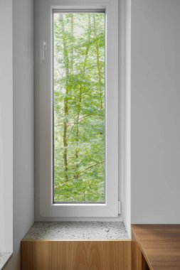 Beyaz ve plastik çerçeveli sakin bir iç mekan ve arka planda ormandan gelen canlı yeşillik. Bir kır evinden doğaya bakış açısı, dikey çekim. Dairenin içindeki elementler
