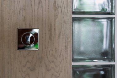 Metal çerçevedeki modern ışık anahtarı, doğal ışığın iç mekanı aydınlatmasını sağlayan cam bloklara bitişik ahşap bir duvara yerleştirilmiştir.