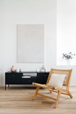 Modern dairede minimalist oturma odasının dikey görüntüsü. Rahat ahşap sandalye ve soyut sanat eserlerinin altında parlak doğal ışıkla aydınlatılmış karanlık bir dolap.