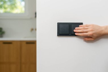 Elektrik kesintisi sırasında ekonomik tüketim enerjisi. Kadın el ile düğmeye basar, beyaz bir fotokopi duvarındaki ışık düğmesini açar. Arka planda ahşap dolaplı mutfak görünüyor.