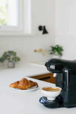 Siyah espresso makinesinin dikey görüntüsü parlak sabah ışığında minimalist bir mutfak tezgahında altın bir kruvasan tabağının yanında kahve demler.