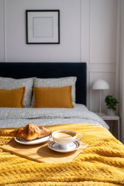 Konforlu otel süitinde yatak ve kahvaltı. Bir tepside kruvasan ve sütlü bir fincan sabah kahvesi düzgünce düzenlenmiş arka planda yumuşak yastıklı yumuşak sarı bir battaniyede oturur.