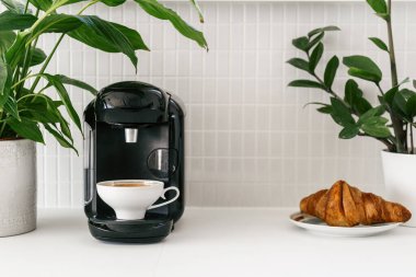 Modern espresso makinesi ve Fransız pastası olan mutfak tezgahı. Tereyağlı kruvasanın yanında beyaz bardakta kapuçino ve seramik kaplarda seramik arka planda canlı yeşil bitkiler olan kahve makinesi.