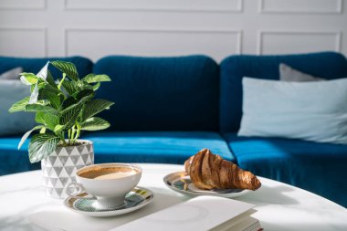 Bir fincan kahve, gevrek kruvasan ve beyaz mermer masa üzerine bir kitap. Oturma odasındaki mavi kanepenin önündeki yiyecek ve içeceğin manzarasını kapat.
