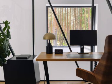 Çağdaş bir ofiste ahşap bir masa, masaüstü monitörü ve pencerelerden görülebilen ağaçların rahatlatıcı görüntüsüne sahip şık bir okuma lambası ve evde huzurlu bir çalışma ortamı yaratılır.