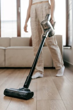 Kadının dikey görüntüsü, iyi aydınlatılmış bir oturma odasındaki ahşap zeminde kablosuz elektrik süpürgesi kullanıyor. Rahat kanepe ve doğal güneş ışığı sıcak bir ev ortamı yaratır.