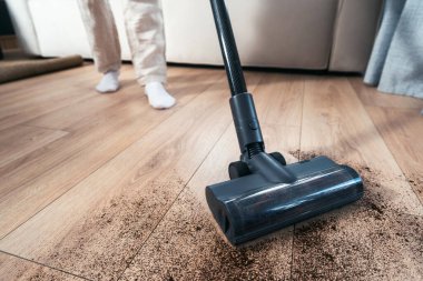 Bir insan, rahat bir oturma odasındaki ahşap zeminden toprak ve tozu temizlemek için elektrikli süpürge kullanıyor. Yakın çekim, fırçaya odaklan