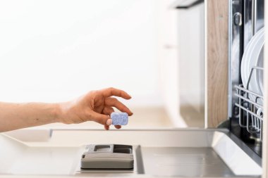 Kadın eli bulaşık makinesine katı bir deterjan ya da tablet koymuş. Kadın bulaşıklarını yıkamak için çevre dostu kapsül kullanıyor. Modern donanımlı mutfak. Ev aletleri, günlük işler.