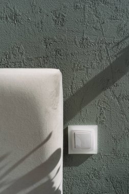 Yatağa yakın yeşil desenli bir duvara monte edilmiş beyaz ışık düğmesinin yakın çekim ve dikey görüntüsü gri yatak başlığıyla. Elektrik teçhizatı ve yumuşak yatak odası mobilyalarına odaklan.